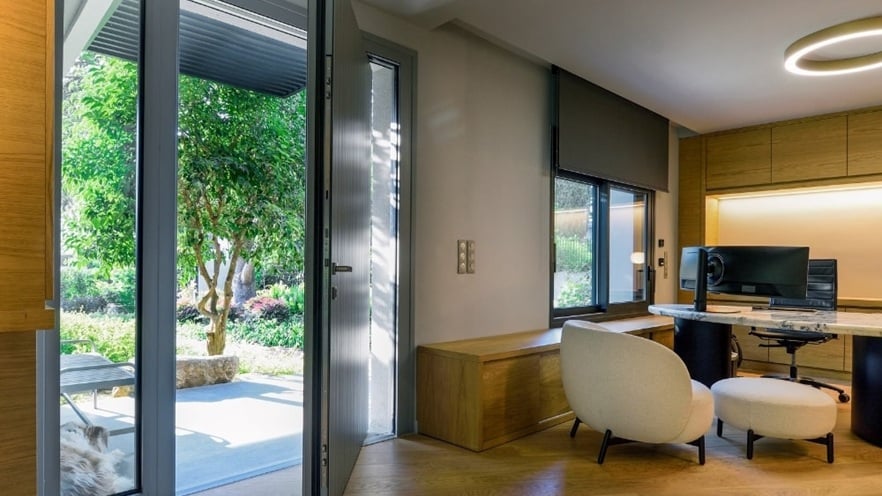 Modern home office with wooden flooring, a large desk and computer, a white chair and ottoman, and an open door leading to a garden; natural light fills the room through glass doors and windows.
