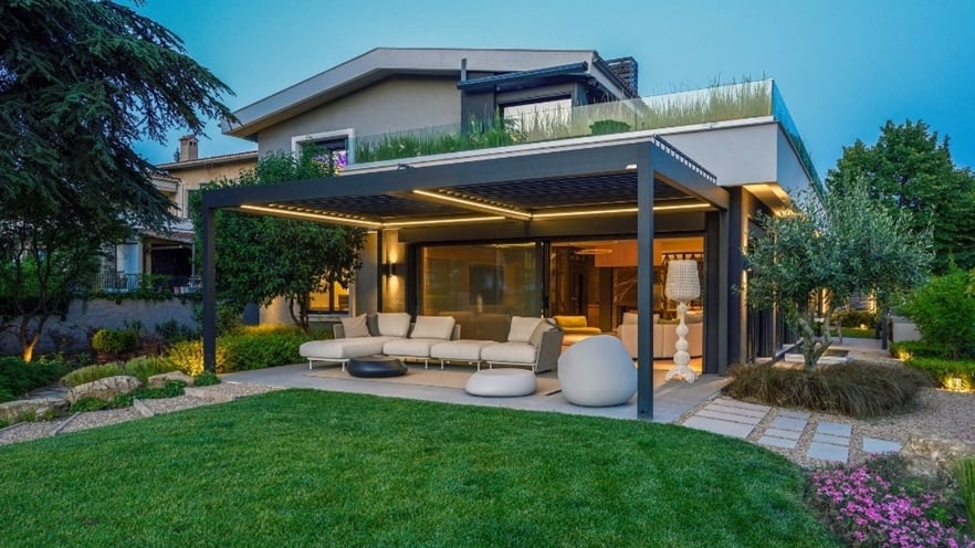 Modern house with a large patio featuring a sectional sofa and round chairs under a pergola with integrated lighting; the surrounding garden has neatly trimmed grass, plants, trees, and flowers.