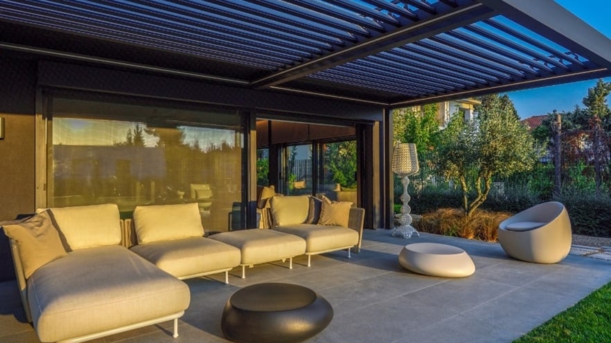 Modern outdoor patio with four beige cushioned seats in an L-shape, two round ottomans, and a black coffee table under a pergola with horizontal louvers; greenery with trees and shrubs in the background.