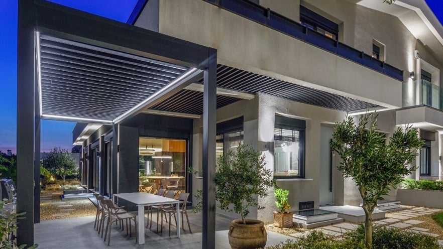 Modern two-story house with a well-lit outdoor dining area under a pergola with integrated lighting; a long table with chairs, large windows, and a landscaped garden with plants and trees, captured in the evening.