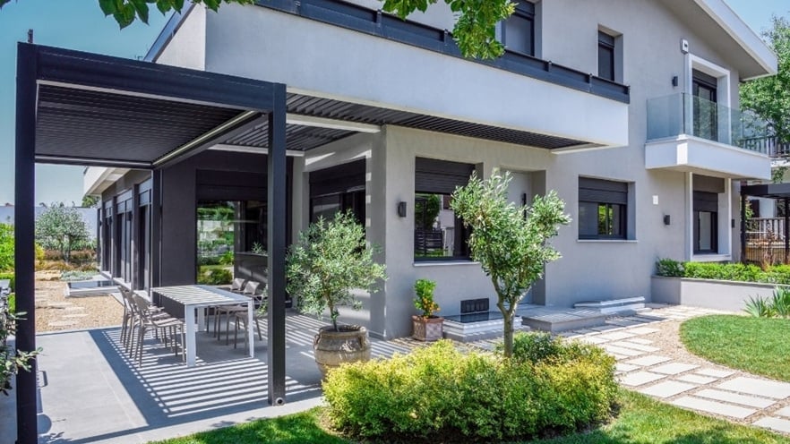 Modern two-story house with a light gray exterior, large windows, and black shutters; outdoor dining area under a pergola with a long table and chairs, a well-maintained garden with bushes, small trees, a stone pathway, and a balcony with glass railings.