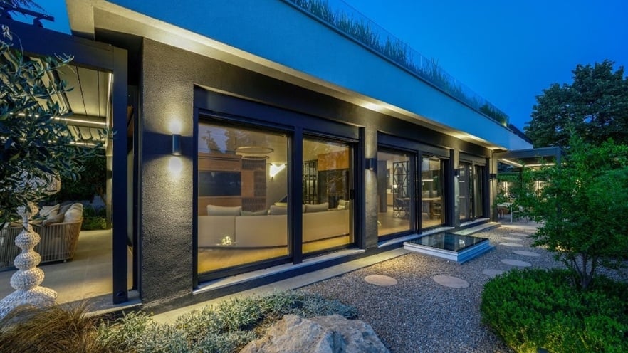 Exterior of a modern house in the evening with large glass windows showing a well-lit interior and a white sofa; exterior walls with outdoor lights, landscaped garden with plants, rocks, gravel pathways, and an outdoor seating area on the left.