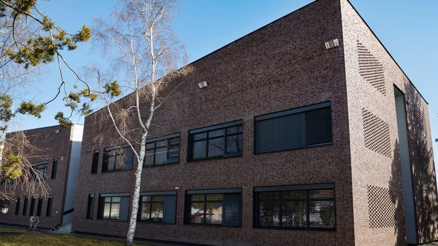 The building has brick walls and large window openings and is surrounded by a courtyard with trees and grass