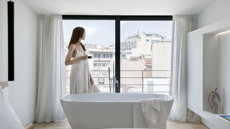 A woman in a white dress looks at the view through the window, in front of the bathtub