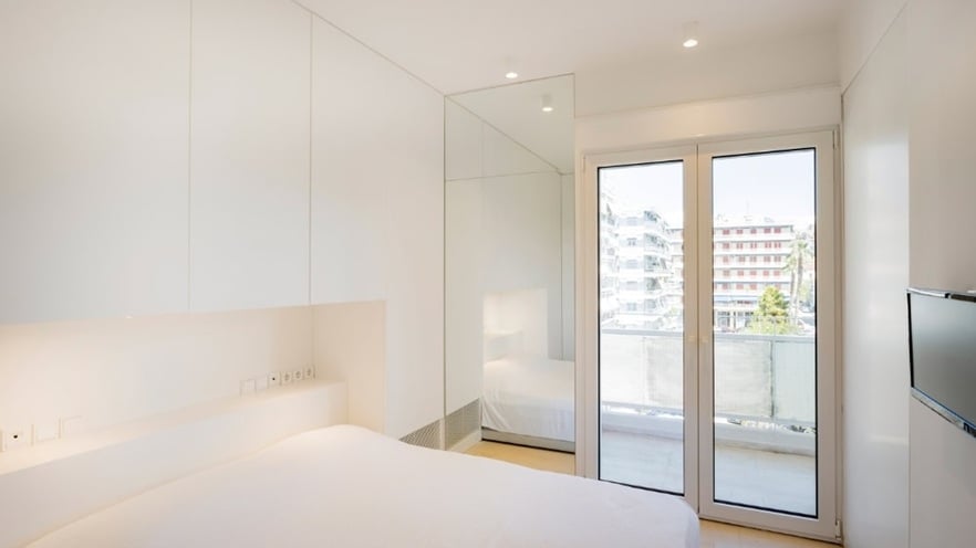 Minimalist bedroom with white walls and furniture, a large bed with white bedding, built-in cabinets, a mirrored wall, sliding glass doors to a balcony, and a wall-mounted TV.
