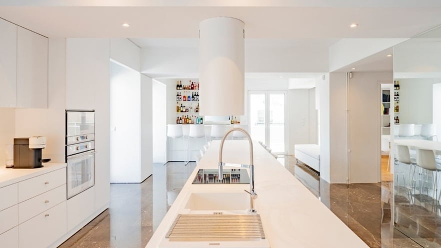 Minimalist kitchen with white cabinets, a central island with built-in sink and cylindrical range hood, shelves of bottles in the background, and bar stools in a naturally lit space.