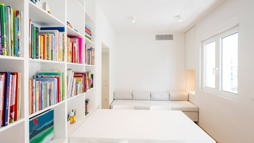 Bright modern room with white walls and furniture, a large bookshelf filled with colorful books, a white table with two chairs, a white sofa, and two windows letting in natural light.