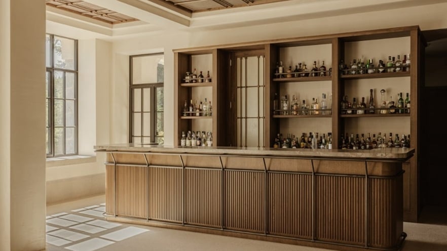 An empty bar area featuring a wooden counter and shelves stocked with various bottles. Natural light streams in through large windows on the left, illuminating the space.