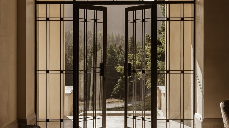 A pair of black-framed double glass doors opened outward, revealing an outdoor view of trees and distant mountains. Natural light streams in, casting shadows on the interior floor.