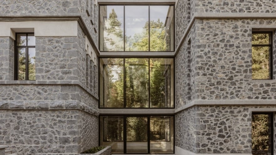 Modern stone building with large glass windows, surrounded by trees and sky reflections.