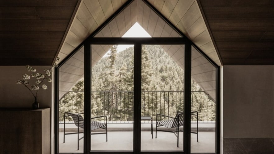 Room with a triangular window opening to a balcony with two chairs, overlooking forested mountains.
