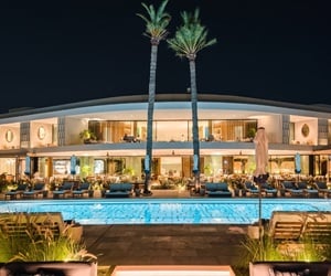 Luxurious modern two-story building at night with a large pool, palm trees, and outdoor seating areas.