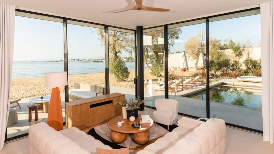 Living room with white sectional sofa, round wooden tables, and beach view through large windows.