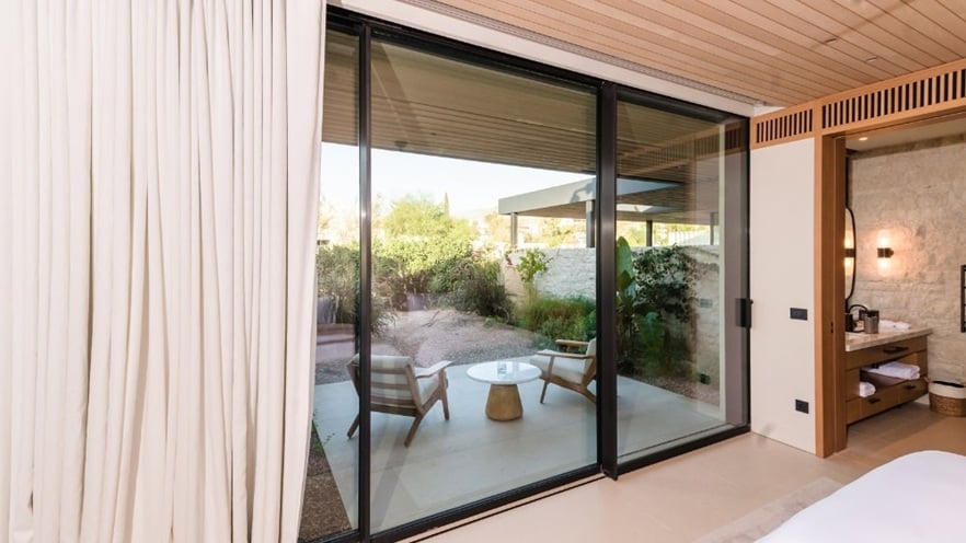 Bedroom with sliding glass door opening to patio with chairs, table, and greenery view.