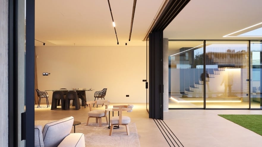 Modern open-concept living space with large aluminium sliding doors opening to a grassy outdoor area. Inside, there is a light-colored sofa, a small wooden coffee table with two stools, and a dining table with four chairs.