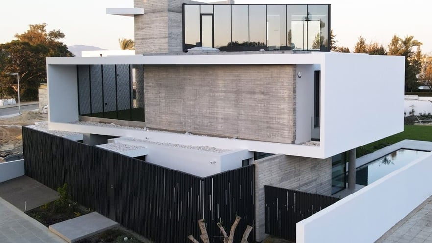 Modern multi-story house with minimalist architecture, featuring large glass windows, concrete and white exterior walls, a black vertical slat fence, and a small front garden. The upper floor extends outward with expansive windows,.
