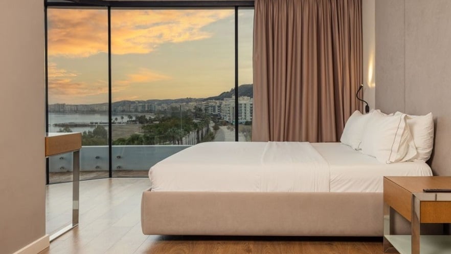 A double bed in front of a large window overlooking the city and the sea