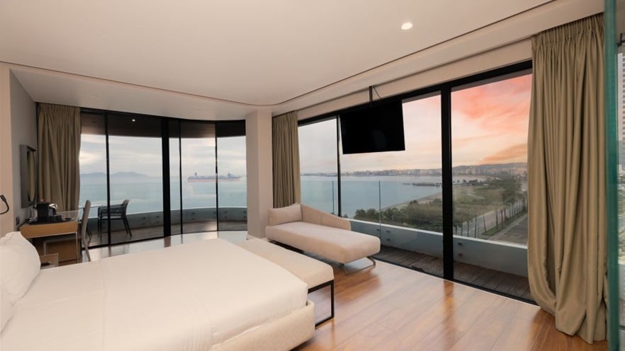The room has large windows overlooking the sea, wooden floors and minimal furniture